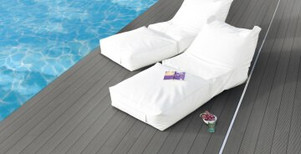 Elegante Poolterrasse mit dunklem WPC-Belag, zwei weißen Lounge-Liegen und ruhiger Wasserreflexion.